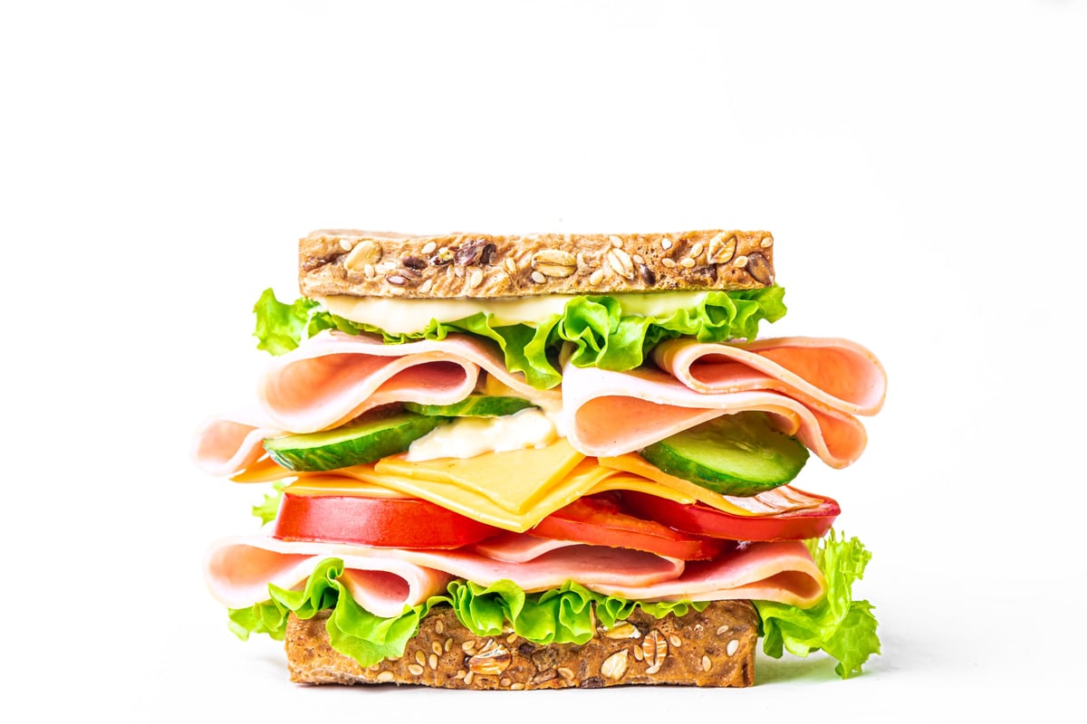 Big layered sandwich with ham, lettuce, cheddar cheese, tomato and cucumber isolated on white background, side view. Healthy lunch sandwich with wholegrain bread, meat, vegetables, cheese and sauce.