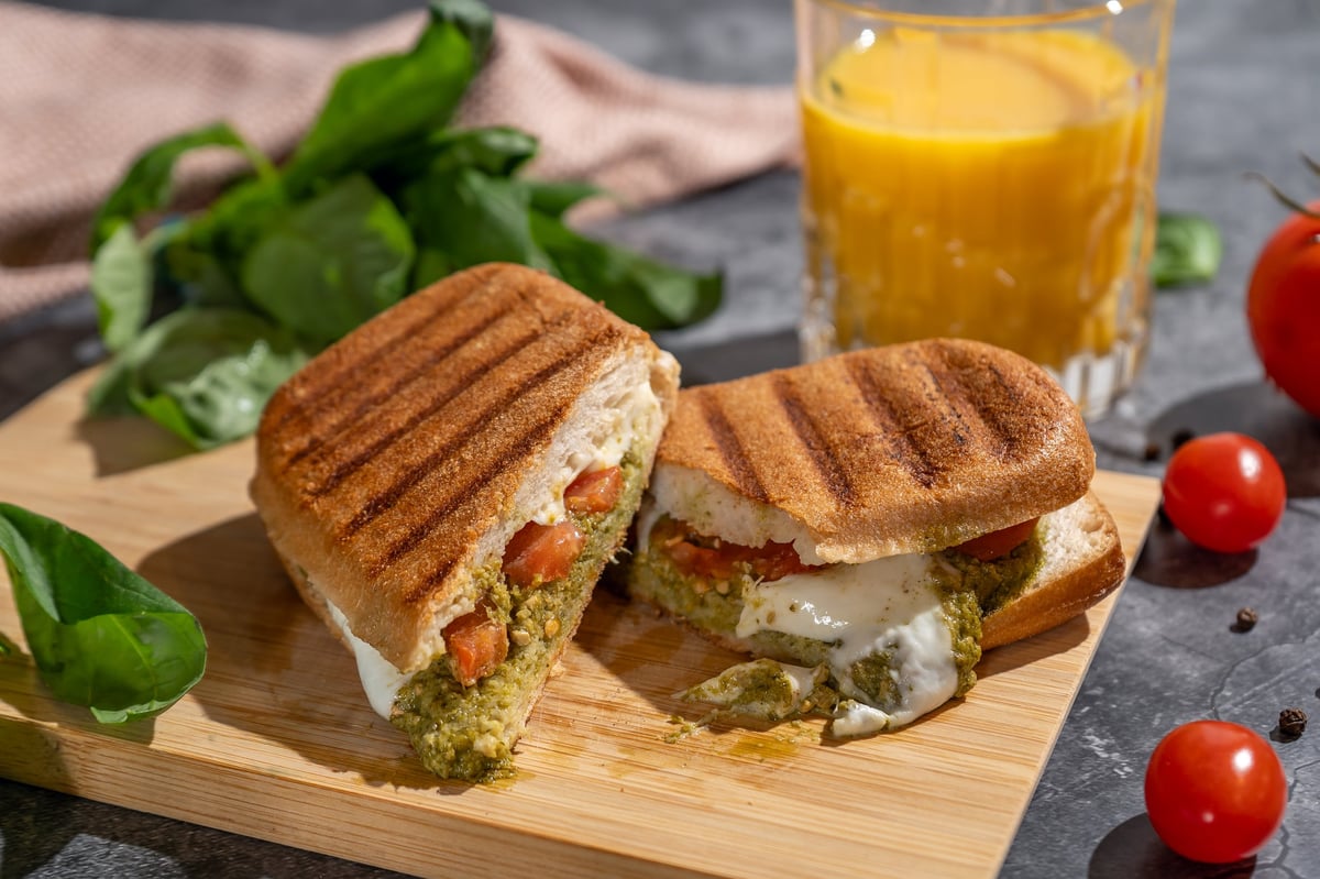 Grilled panini sandwich with pesto, mozzarella, tomato, and basil on a wooden board.