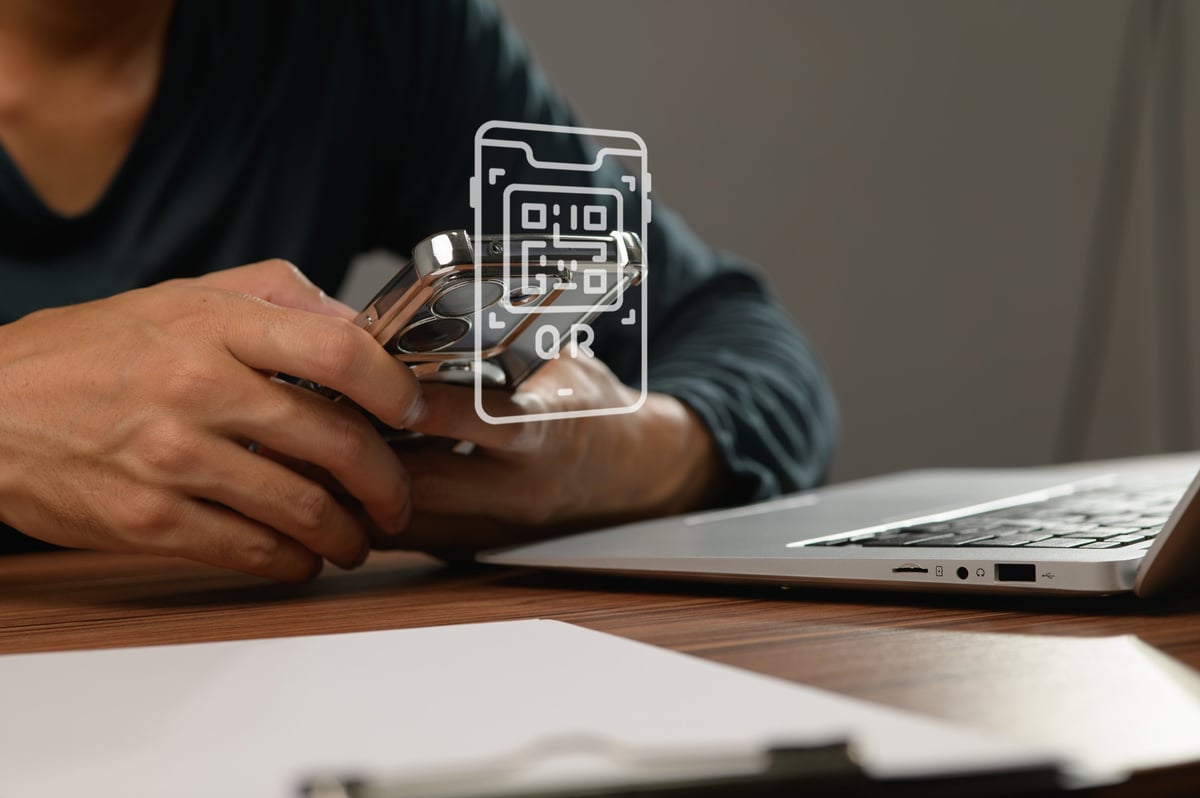 A close-up view shows a person holding a smartphone with a QR code overlay, while sitting at a wooden desk with a laptop. Ideal for technology themes.