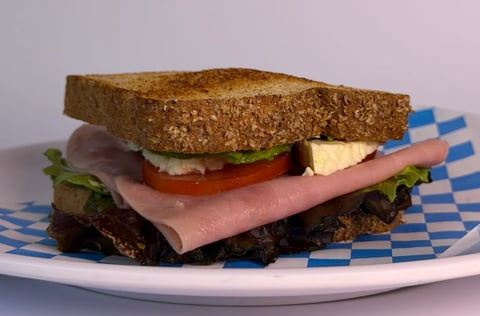 A multi-layered sandwich with whole wheat bread, ham, cheese, tomato, and lettuce on a checkered paper-lined plate against a gray background.