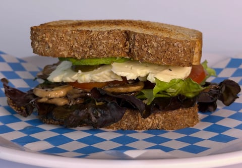 Stacked sandwich with whole grain bread, lettuce, tomato, cheese, and grilled eggplant on a blue and white checkered paper