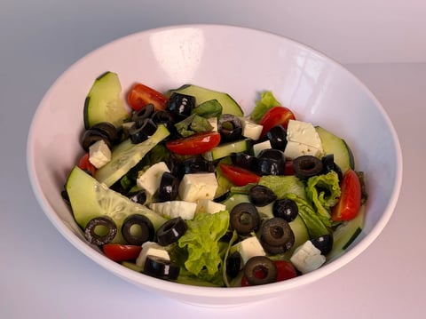 White bowl containing a fresh salad with sliced cucumbers, red peppers, black olives, white cheese cubes, and green lettuce.