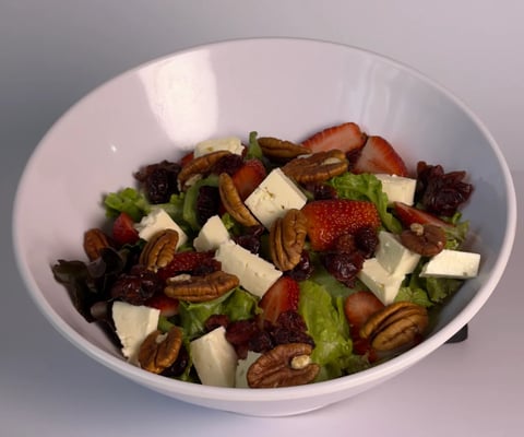 A white bowl containing a colorful salad with mixed greens, strawberries, white cheese cubes, pecans, and dried cranberries.