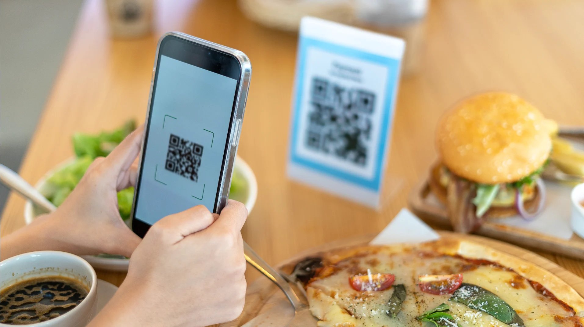 Hand holding smartphone displaying QR code at restaurant table with food and QR code signs