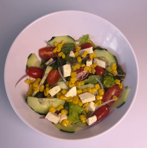 Fresh salad bowl with cucumber slices, tomatoes, corn, feta cheese, and greens on a pink plate