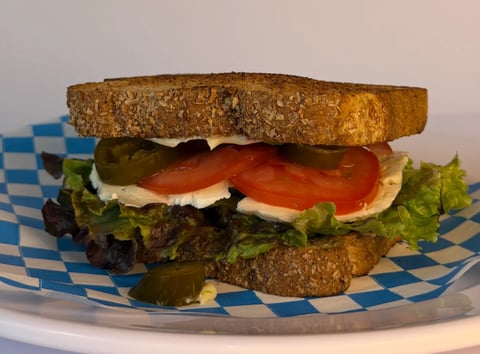 A sandwich with toasted whole grain bread, lettuce, tomato slices, and mayo on a blue and white checkered paper wrapper.